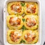 A white baking dish with six baked polenta squares, each topped with tomato sauce, melted cheese, and herbs, garnished with fresh parsley. Nearby, a beige cloth and parsley sprigs create a cozy scene reminiscent of chicken and mashed potatoes comfort.
