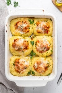 A white baking dish with six baked polenta squares, each topped with tomato sauce, melted cheese, and herbs, garnished with fresh parsley. Nearby, a beige cloth and parsley sprigs create a cozy scene reminiscent of chicken and mashed potatoes comfort.
