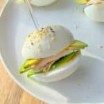 A hand holds a toothpick, securing an Egg Sandwich made with egg halves as buns, filled with turkey, cheese, lettuce, and avocado, garnished with sesame seeds—all served on a white plate.