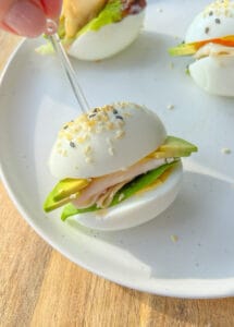 A hand holds a toothpick, securing an Egg Sandwich made with egg halves as buns, filled with turkey, cheese, lettuce, and avocado, garnished with sesame seeds—all served on a white plate.