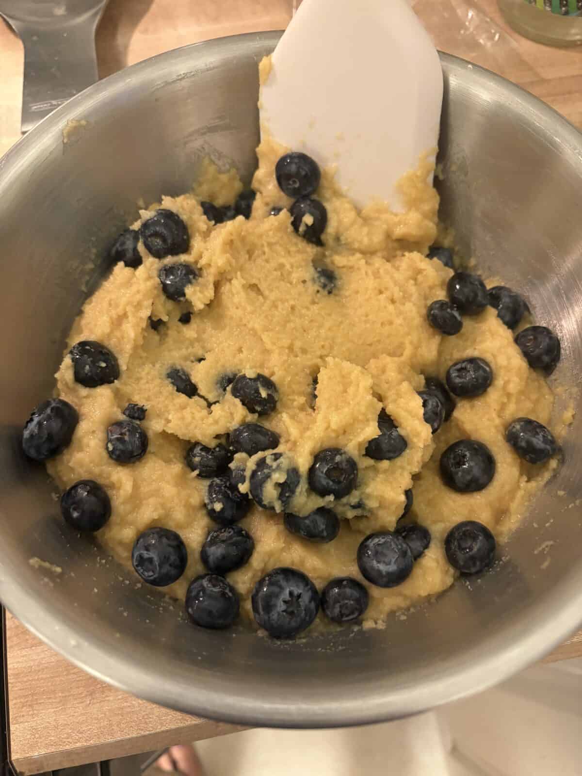A metal mixing bowl containing thick yellow batter for Almond Flour Blueberry Muffins, with fresh blueberries being stirred in with a white spatula.