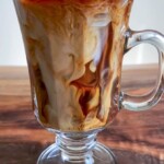 A glass mug filled with Iced Coffee with Vanilla, swirling with cream, sits on a wooden surface. The drink displays marbled patterns as the vanilla-scented cream mixes with the coffee over ice.