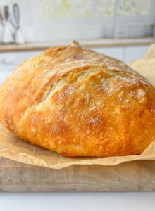 A golden-brown loaf of crusty bread sits on parchment paper atop a wooden board in a bright kitchen, ready to be paired with creamy whipped Brie.