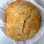 A round, golden-brown loaf of No Knead Cheddar Bread with a rustic crack on top rests on a white cloth, promising a crusty bite and cheesy aroma.