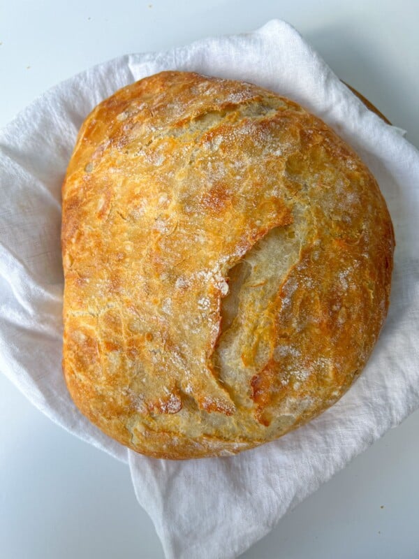 A round, golden-brown loaf of No Knead Cheddar Bread with a rustic crack on top rests on a white cloth, promising a crusty bite and cheesy aroma.