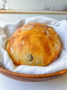 A golden, freshly-baked loaf of bread with a glossy crust rests on a white cloth inside a wooden bowl, set on a white surface in a bright kitchen—perfect for pairing with creamy whipped Brie.