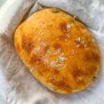 A freshly baked, golden-brown No Knead Olive Bread with a shiny crust and visible olives rests on a white linen cloth inside a basket.