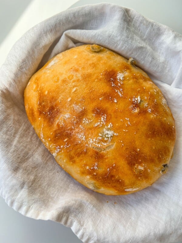 A freshly baked, golden-brown No Knead Olive Bread with a shiny crust and visible olives rests on a white linen cloth inside a basket.