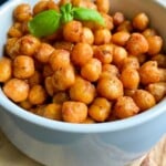 A white bowl filled with air fryer chickpeas, a high-protein snack, garnished with a sprig of fresh basil sits on a wooden board.