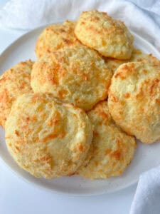A plate of golden, freshly baked Cheese Buns stacked on top of each other, with a white cloth in the background.