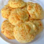 A plate of golden, fluffy cheese buns piled on top of each other, with a light, crispy exterior and visible bits of melted cheese on a white plate.