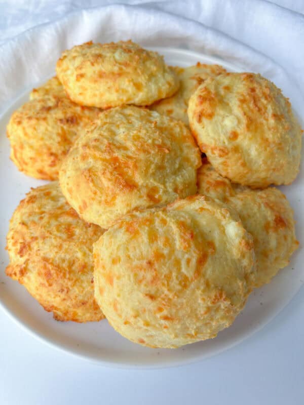 A plate of golden, fluffy cheese buns piled on top of each other, with a light, crispy exterior and visible bits of melted cheese on a white plate.
