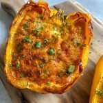 A yellow bell pepper half, filled with a golden-brown cheesy Breakfast Stuffed Peppers filling and garnished with chopped herbs, sits on a wooden cutting board.
