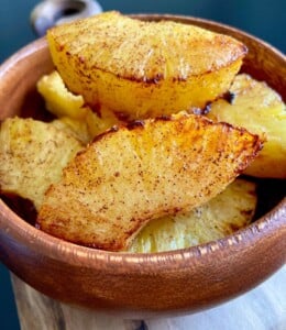 A wooden bowl filled with golden brown, grilled pineapple slices dusted with cinnamon, placed on a wooden surface with a dark background.