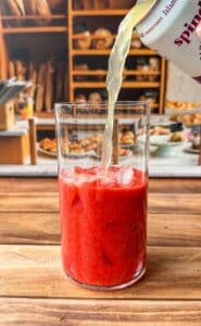 A clear glass filled with red, icy strawberry puree is being topped with a light-colored sparkling drink, on a wooden table with a bakery display blurred in the background.