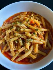 A white bowl filled with Fasolakia—braised green beans in a tomato-based sauce—garnished with herbs and small vegetable pieces, set against a dark background.
