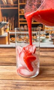 A glass filled with ice cubes is being filled with thick red juice poured from a pitcher. In the background, there are shelves with bread and pastries, giving a cozy bakery atmosphere.