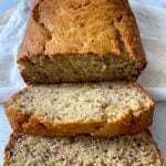 A loaf of Gluten Free Banana Bread with a golden-brown crust, partially sliced to reveal its moist, speckled interior. Two slices are laid in front on a white surface with a white cloth underneath.