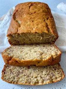 A loaf of Gluten Free Banana Bread with a golden-brown crust, partially sliced to reveal its moist, speckled interior. Two slices are laid in front on a white surface with a white cloth underneath.