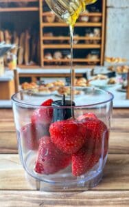 A close-up of fresh strawberries in a food processor bowl as honey is being poured over them, with a bakery display blurred in the background.