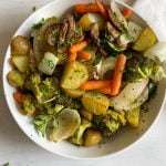 A white bowl filled with oven roasted vegetables—carrots, broccoli, potatoes, onions, and mushrooms—garnished with chopped herbs and placed on a light-colored surface.