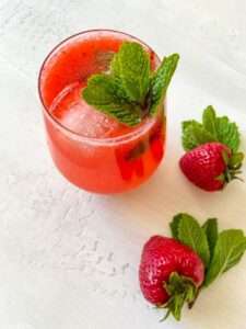 A glass of Strawberry Mojito with ice, garnished with fresh mint leaves. Two whole strawberries and mint leaves are placed beside the glass on a light surface.