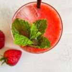 A top view of a glass filled with a vibrant red Strawberry Mojito, ice cubes, and fresh mint leaves, with a black straw. Two strawberries rest on the white surface beside the glass.