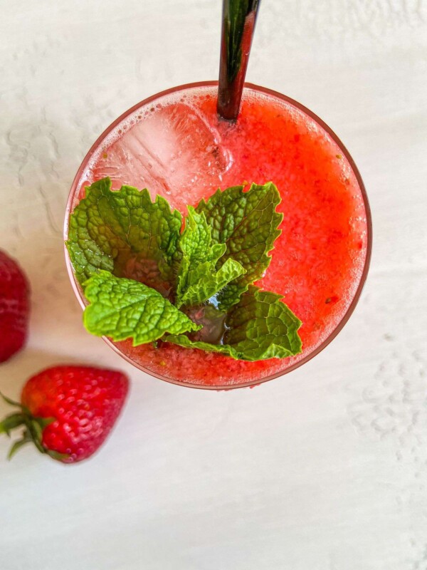 A top view of a glass filled with a vibrant red Strawberry Mojito, ice cubes, and fresh mint leaves, with a black straw. Two strawberries rest on the white surface beside the glass.