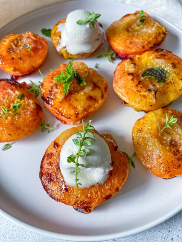 A white plate with Air Fryer Peaches—grilled halves, some topped with a scoop of vanilla ice cream and fresh herb sprigs as garnish.