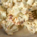 Close-up of a bowl of creamy roasted cauliflower on a beige plate. A fork is holding a piece of cauliflower, displaying a golden-brown edge, with sauce and herbs visible. The dish is set on a light-colored surface.