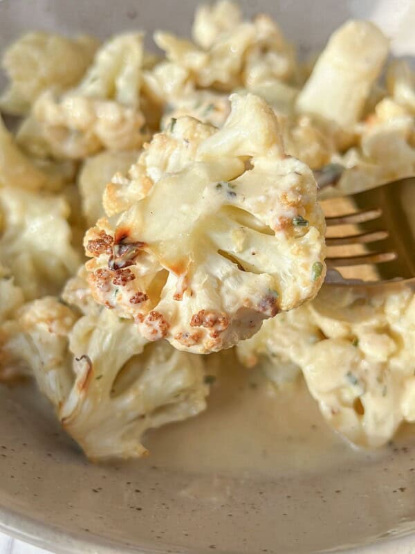 Close-up of a bowl of creamy roasted cauliflower on a beige plate. A fork is holding a piece of cauliflower, displaying a golden-brown edge, with sauce and herbs visible. The dish is set on a light-colored surface.