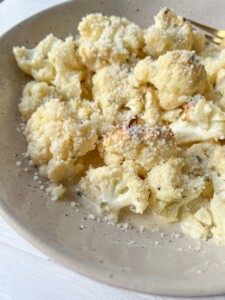 A bowl of roasted cauliflower florets topped with grated cheese, served on a beige ceramic plate. A fork is partially visible on the side. The dish is set against a light background.