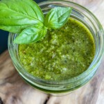A small glass jar filled with vibrant Basil Walnut Pesto, garnished with two fresh basil leaves, sits on a wooden surface.
