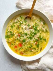 A bowl of The Best Chicken Soup Ever, garnished with chopped parsley, features shredded chicken, rice, carrots, and celery in a clear broth, with a wooden spoon resting inside the bowl.