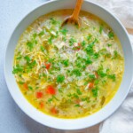 A bowl of The Best Chicken Soup Ever, filled with chicken, rice, chopped carrots, celery, and parsley, is served with a wooden spoon on a white napkin and light-colored surface.