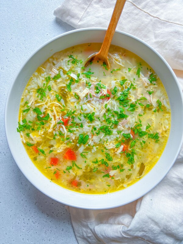 A bowl of The Best Chicken Soup Ever, filled with chicken, rice, chopped carrots, celery, and parsley, is served with a wooden spoon on a white napkin and light-colored surface.