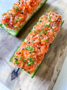 Two cucumber halves topped with diced salmon, herbs, and sesame seeds are arranged on a wooden board—an elegant twist on classic Egg Salad Cucumber Boats, garnished with chopped greens and colorful seasoning.