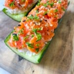 Enjoy Cucumber Salmon Boats—two cucumber boats filled with white rice and topped with diced salmon, sprinkled with black and white sesame seeds and chopped parsley, served on a wooden board.