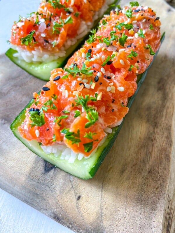 Enjoy Cucumber Salmon Boats—two cucumber boats filled with white rice and topped with diced salmon, sprinkled with black and white sesame seeds and chopped parsley, served on a wooden board.