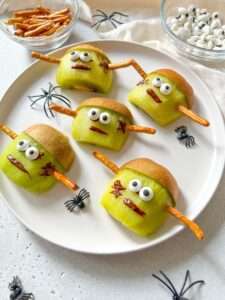 Five Halloween Frankenstein Kiwis, each with candy eyes, pretzel stick arms, and red icing scars, are arranged as spooky faces on a white plate surrounded by plastic spiders, with bowls of pretzels and candy eyes nearby.