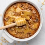 A round baked oatmeal dish topped with walnuts and drizzled with honey. A honey dipper rests on top, and oats are scattered on the white surface surrounding the dish.
