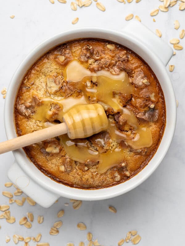 A round baked oatmeal dish topped with walnuts and drizzled with honey. A honey dipper rests on top, and oats are scattered on the white surface surrounding the dish.