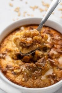 A spoonful of warm bread pudding with walnuts and honey, held above a white ramekin filled with more pudding. The dessert has a golden-brown crust and is drizzled with honey, showing a moist, flavorful texture.