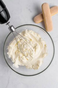A mixing bowl filled with creamy, whipped mascarpone sits on a marble countertop. A hand mixer with a whisk attachment is placed above the bowl. Two ladyfinger cookies are positioned beside the bowl.