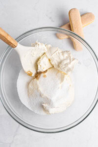 A glass bowl contains cream cheese, mascarpone, sugar, and vanilla extract being mixed with a spatula. Two ladyfinger cookies are placed beside the bowl on a light surface.