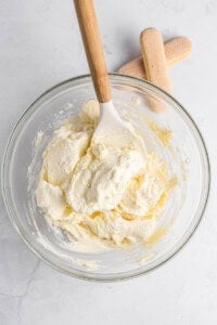 A glass bowl filled with creamy tiramisu mixture being stirred with a wooden spoon, set on a marble surface. Two ladyfinger cookies are placed beside the bowl.