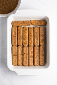A rectangular white dish filled with neatly arranged ladyfingers soaking in coffee, with one missing from the top row. A bowl with remaining coffee is partially visible in the top left corner, on a white marble surface.