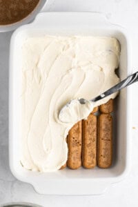 A white baking dish with a layer of ladyfingers partially covered by a thick layer of creamy filling being spread with a spoon. A small bowl with cocoa powder is visible in the background on a marble surface.