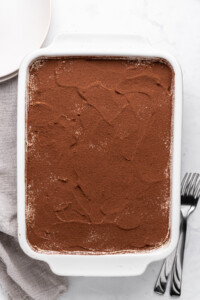 A rectangular dish of tiramisu, topped with a layer of cocoa powder. The dish is on a light countertop with a gray napkin, a plate, and two forks beside it.