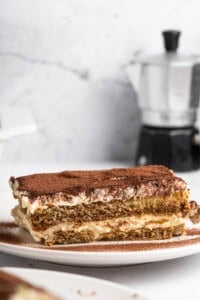 A slice of tiramisu on a white plate, with layers of coffee-soaked ladyfingers and creamy mascarpone, dusted with cocoa powder. A blurred coffee pot is in the background.